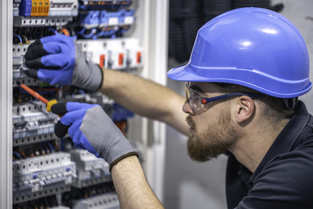 Eletricista com capacete, óculos e luvas de proteção realizando manutenção em painel elétrico, representando a segurança no trabalho do curso NR 10.